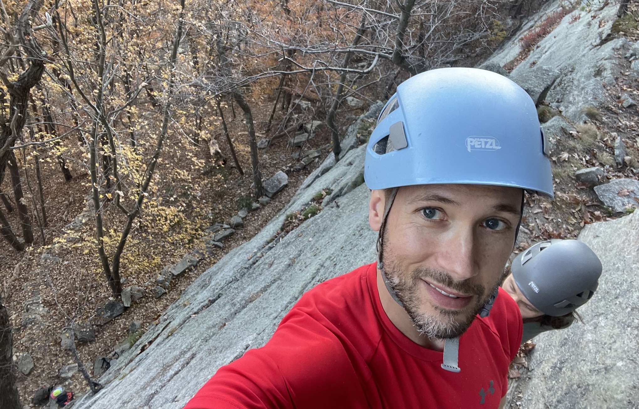 Selfie, smiling, wearing blue climbing helmet, standing on a rocky ledge with fall leaves and bare trees far below.