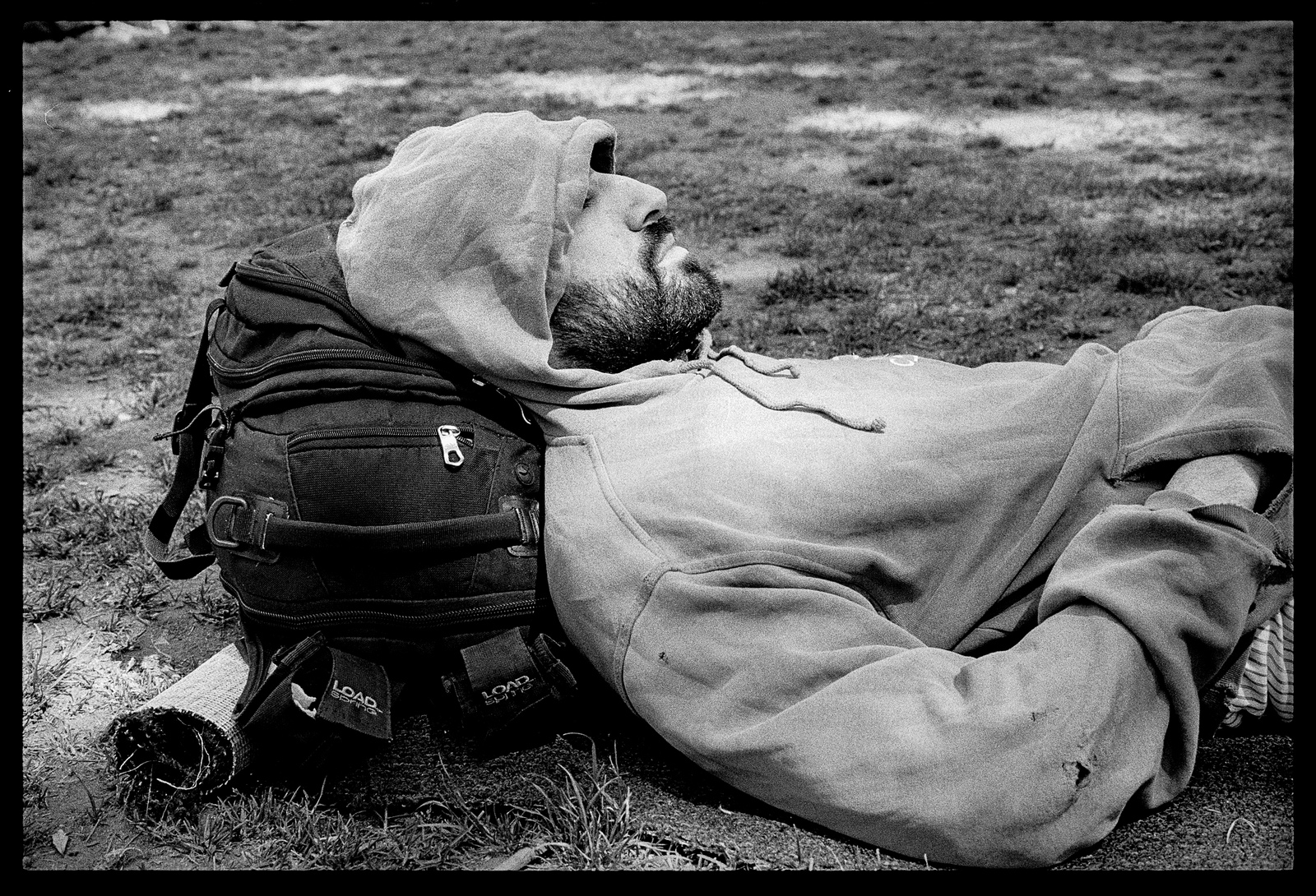 Image description: A young man with a scruffy beard sleeps on the grass in dappled sunlight. His hood is drawn partly over his head and his hands are in the pockets of his hoodie. His head rests on a backpack.