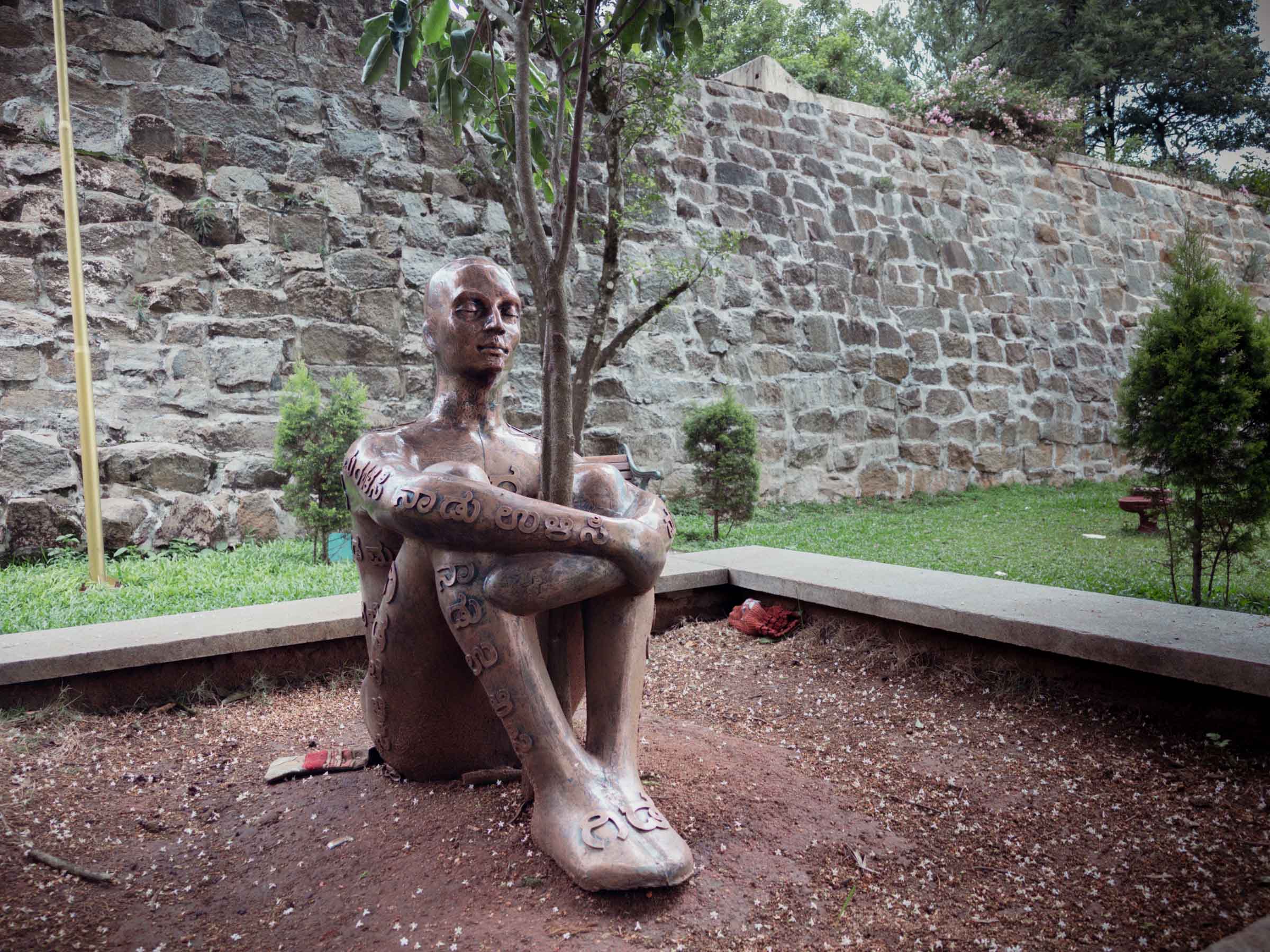 A bronze statue of a seated figure embracing a sapling, its limbs inscribed with text