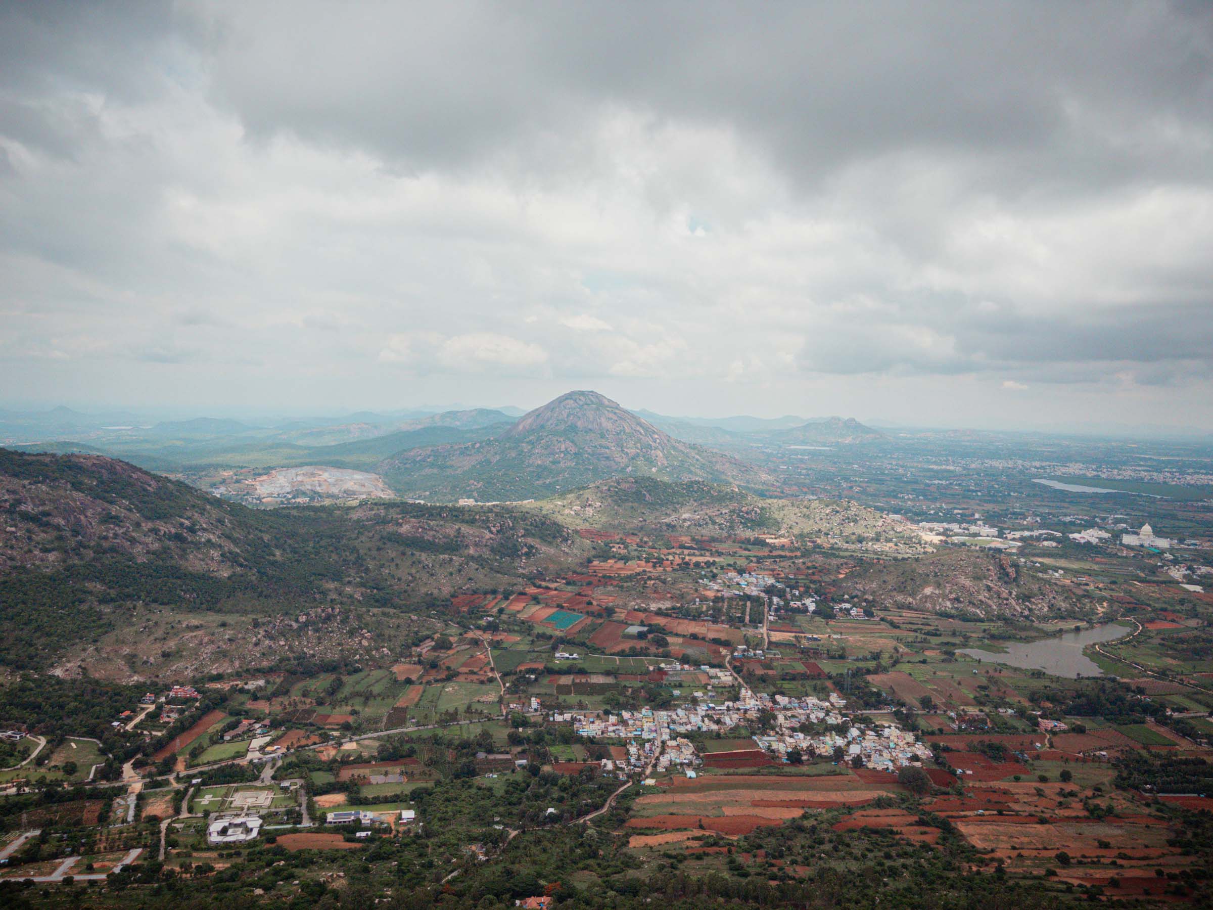 A view from the hills over green slopes and red farmland, with a conical peak in the distance