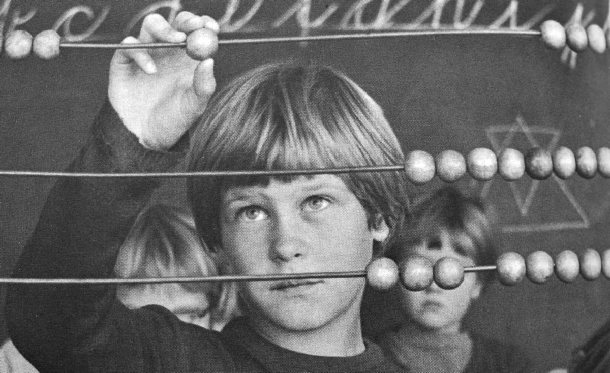 Description: Black and white photo. A boy stands behind a very large abacus that fills the image. He looks up at the ball he is moving on one of the abacus's wires, above his eye-level. Behind him are two schoolchildren and a chalkboard with indistinct writing and diagrams.