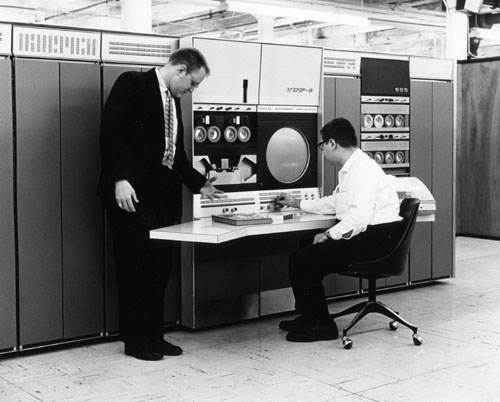 Black and white photo of a DEC PDP-6, a mainframe computer the height of a refrigerator and the width of four of them. Its central cabinet has an oscilloscope, four dials, and a reel-to-reel tape. A man stands in front of it in a suit looking down at the console, while a man in shirtsleeves sits at the console with his hands at the controls.