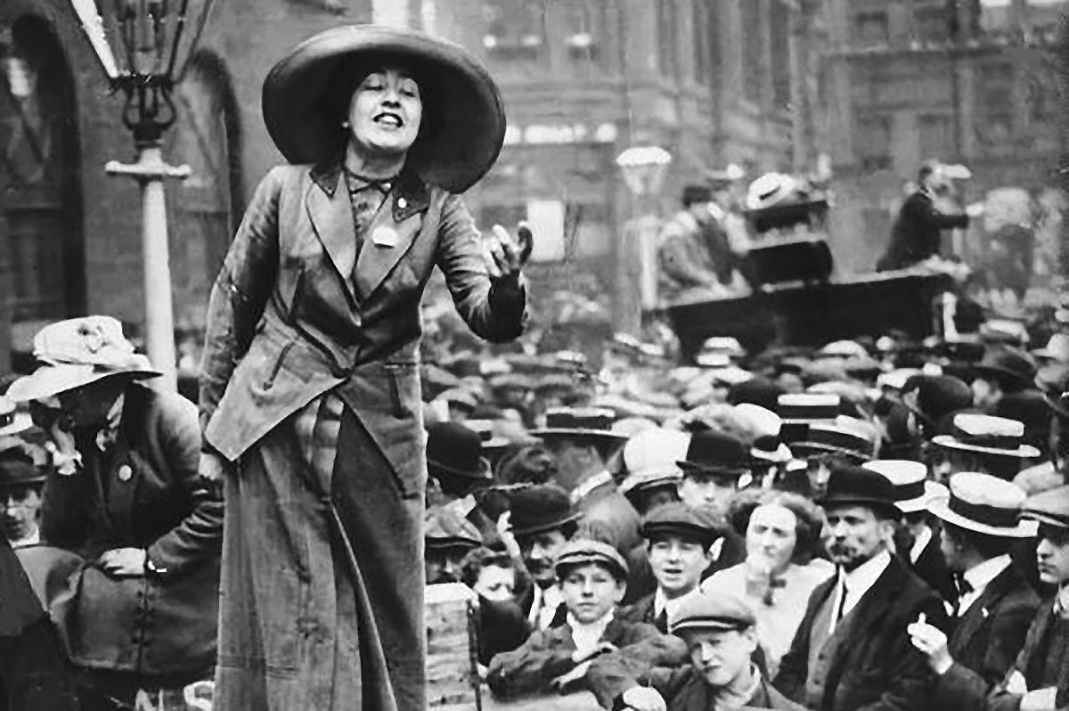 Black and white photo of Sylvia Pankhurst addressing crowd of men in Trafalger Square. She is wearing a large old-fashioned hat and her hand is raised in gesture.