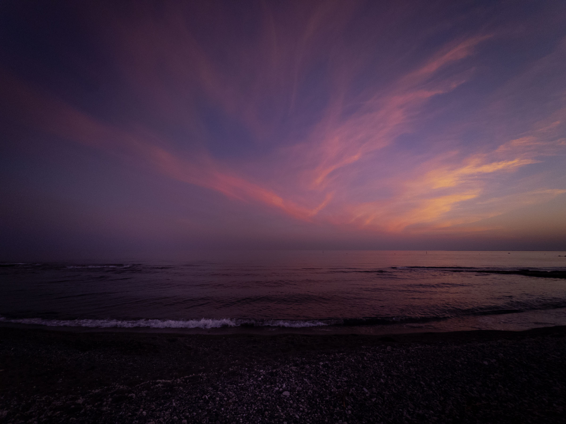 Pink and orange streaks of cloud over the sea at dusk, with waves breaking on a pebble beach