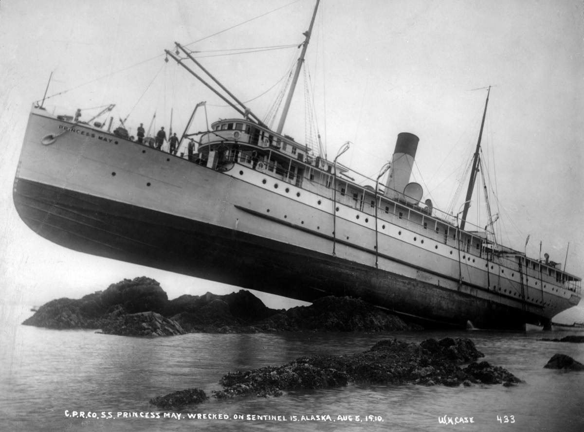 Black and white photo. The steamship Princess May ran aground in 1910 on rocks near the north end of Sentinel Island. It was high tide and the momentum of the ship forced it well up onto the rocks, with the bow jutting upward at an angle of 23 degrees.