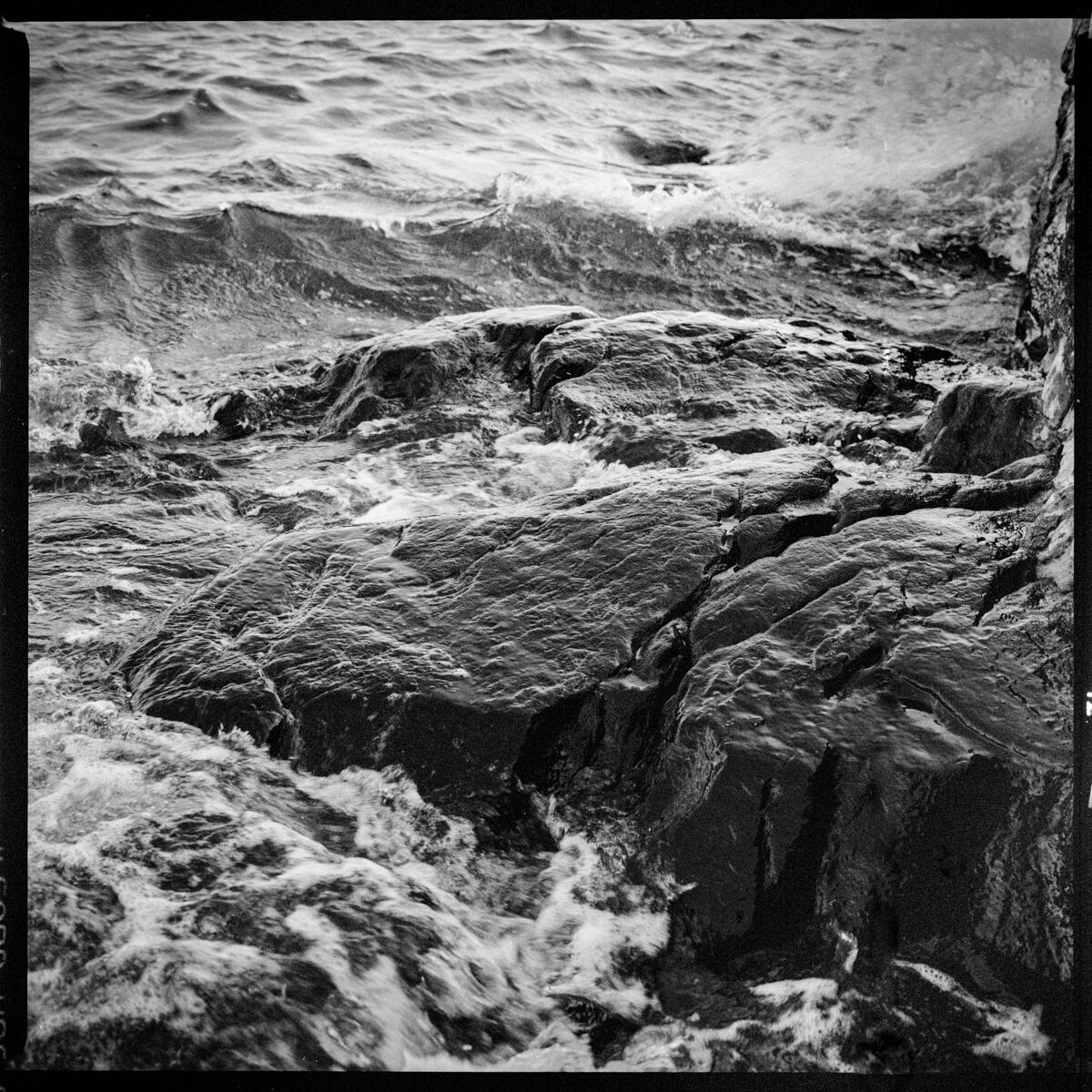 Image description: black and white photograph of a black rock surrounded by waves, seen from above, with surf spraying over one edge of the rock.