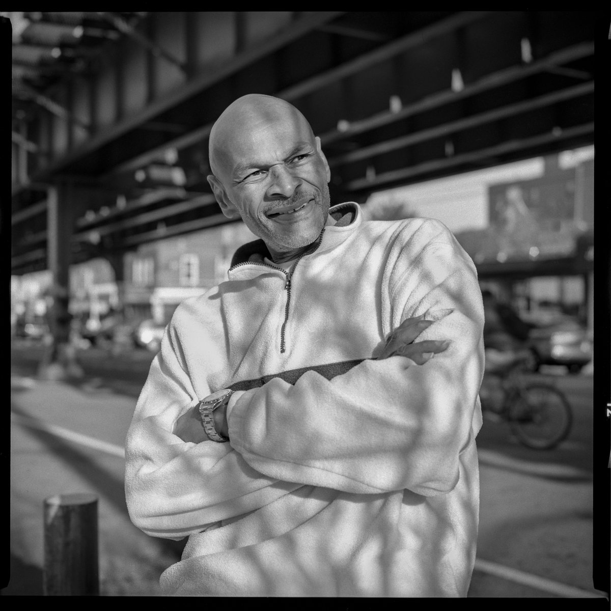 Image description: John, a slim middle-aged black man with a shaved head, looks to the side and smiles. He sits beneath an elevated train bridge in an urban neighborhood. Shadows dapple him.