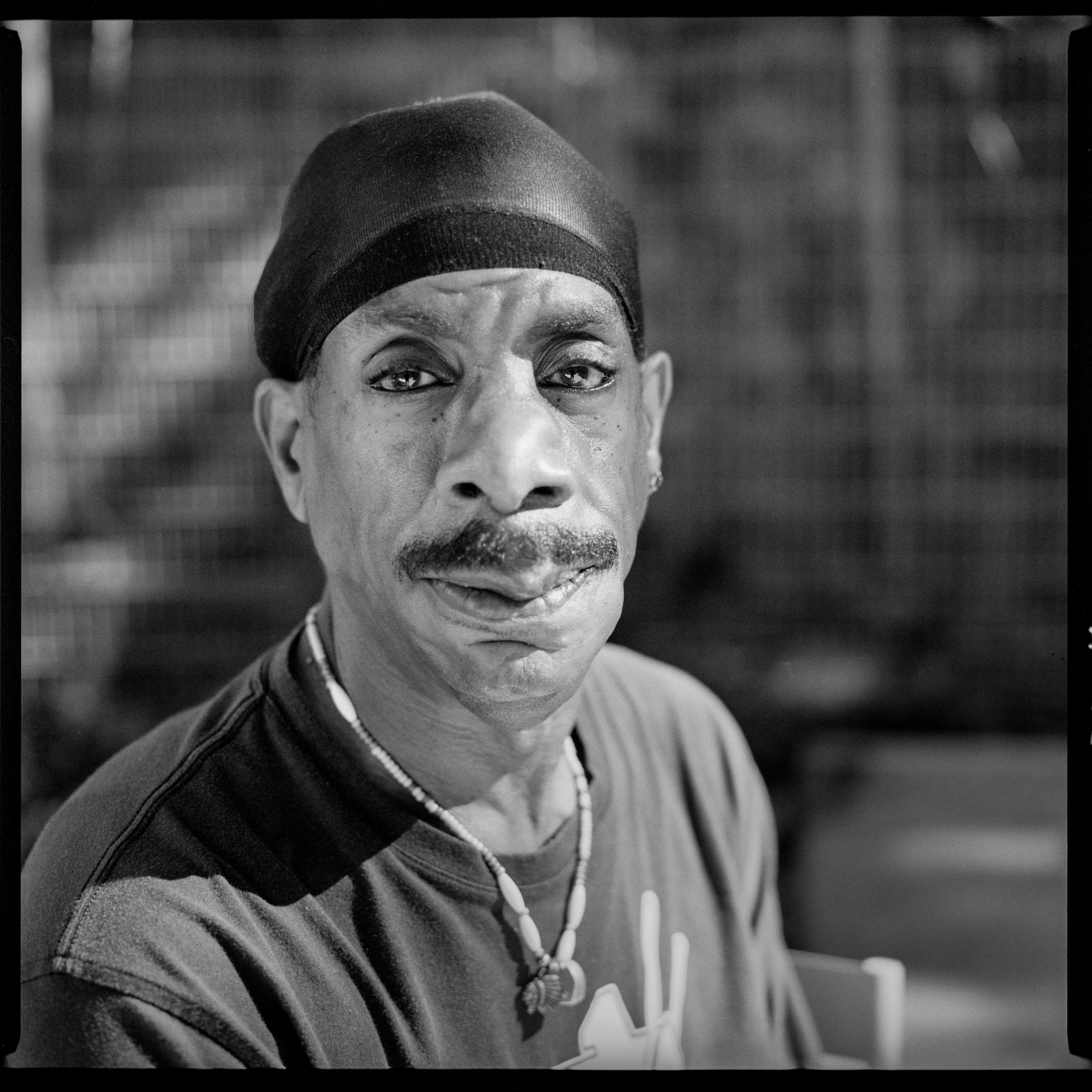 Image description: Black and white photograph of a black man with mustache, wearing a do rag, a T-shirt, and a bead necklace. He is looking into the camera, his eyes are lined with eyeshadow. His nose and jaw are asymmetrical as if from an injury.