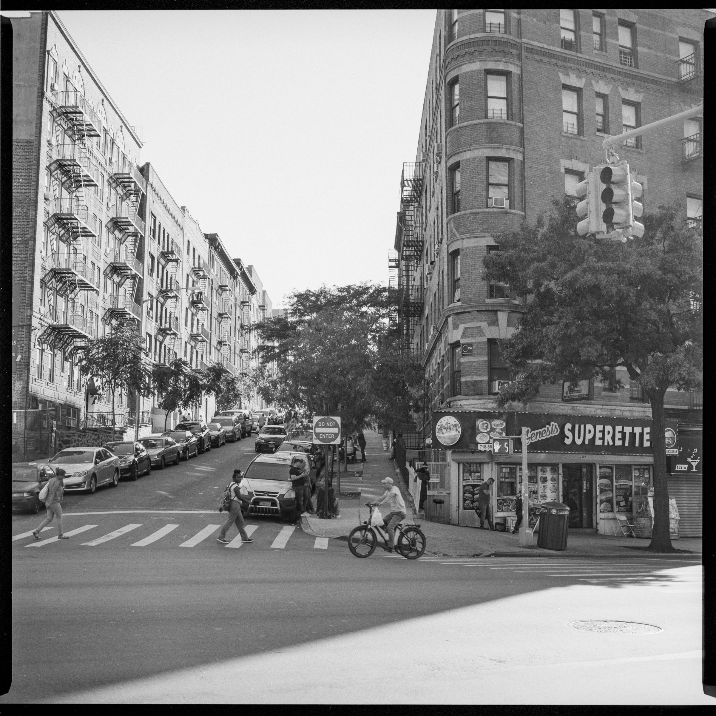 Image description: Black and white photograph of an urban street corner. A bodega storefront has the sign “Genesis Superette”. A bicycle and two pedestrians are crossing in front of the camera. A steep hill lined with apartment buildings recedes in the background.