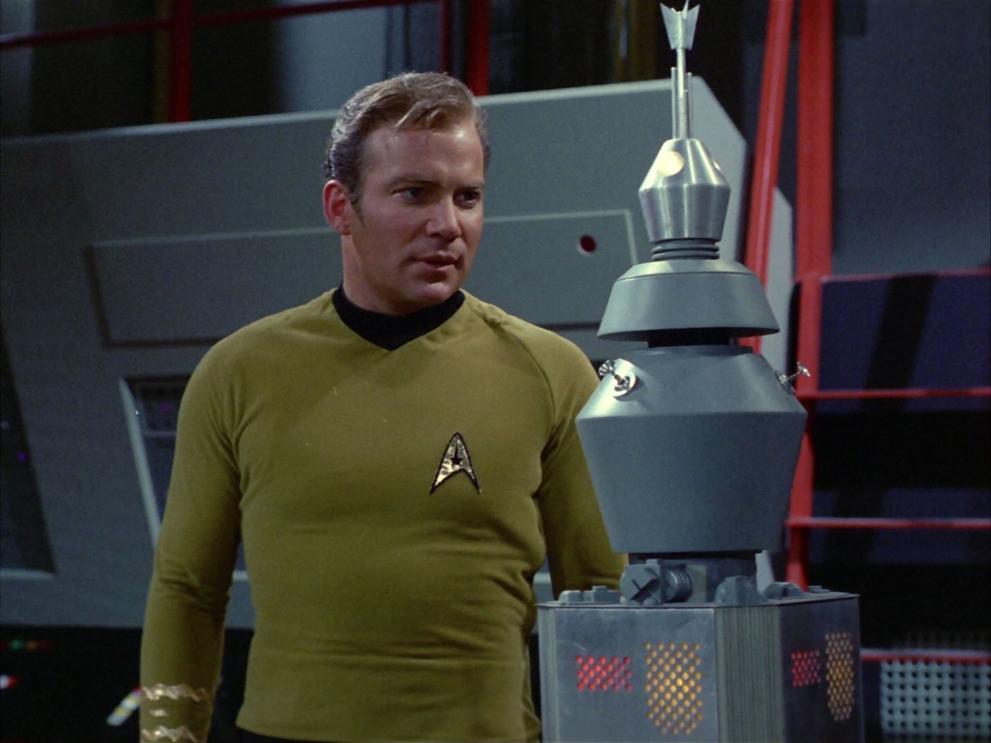 Captain Kirk from Star Trek the original series. He’s looking angrily at a computer, which is in the shape of a gray cylinder with a few lights, knobs, and antennae.