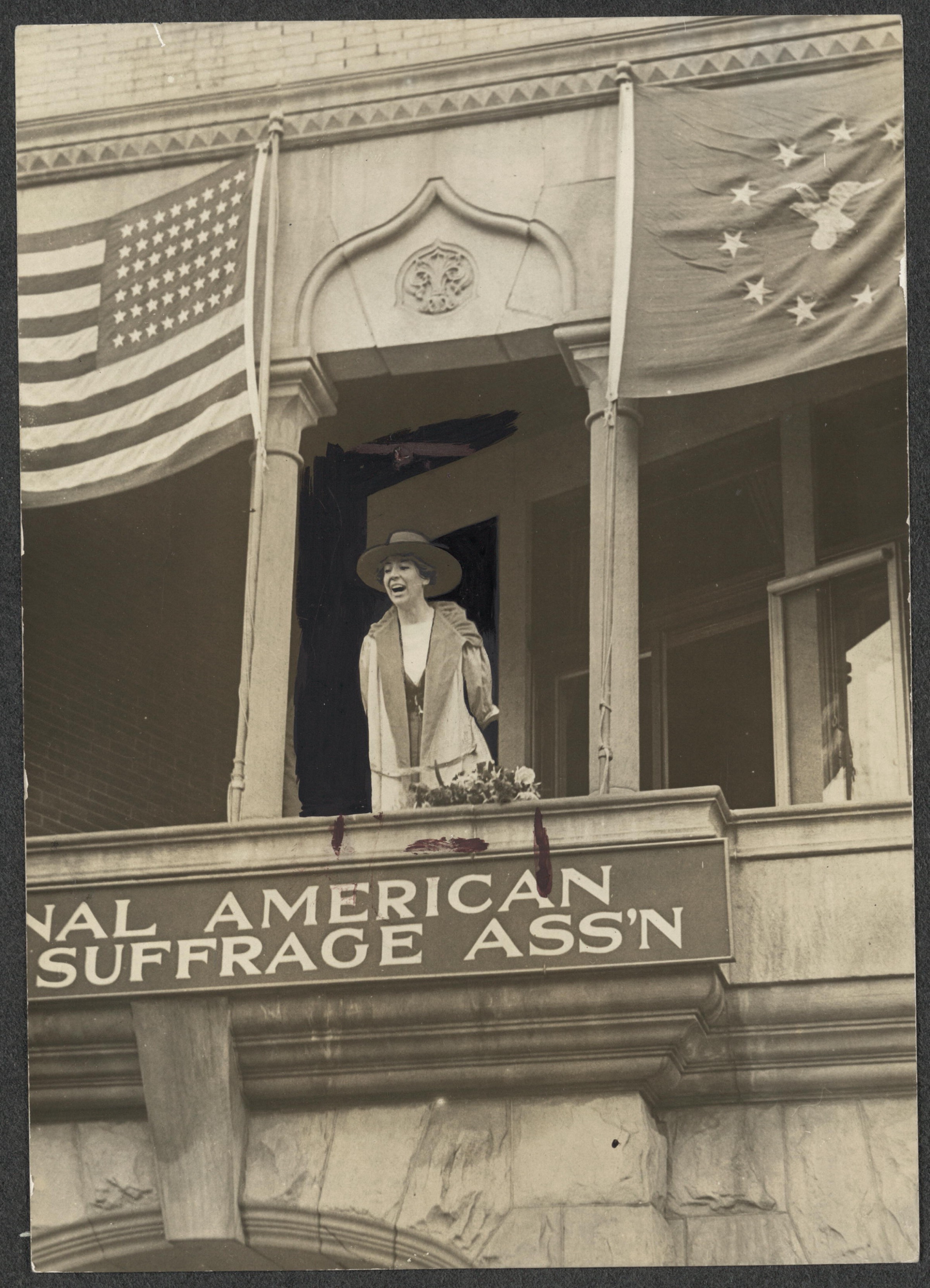 Black and white 1917 photo of a woman speaking from a balcony. A sign saying “National American Woman Suffrage Association” is partly visible beneath her.