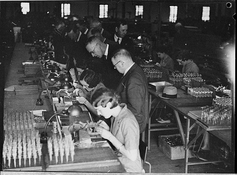 A supervisor watches workers assembling small parts at a long factory bench, circa 1936.