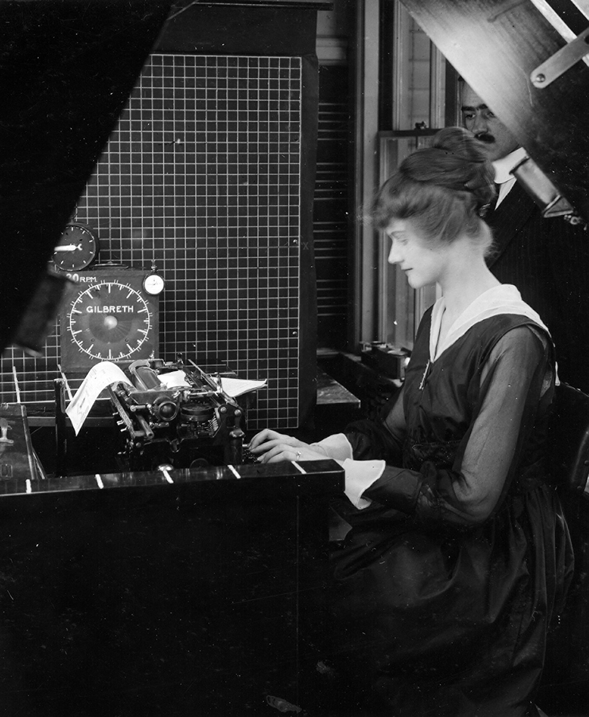 A woman operates a typewriter while being filmed, with a large clock labeled 'Gilbreth' visible beside her.