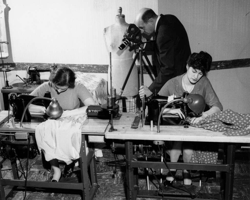 A man films two women working at sewing machines with a motion picture camera on a tripod, in a time and motion study.