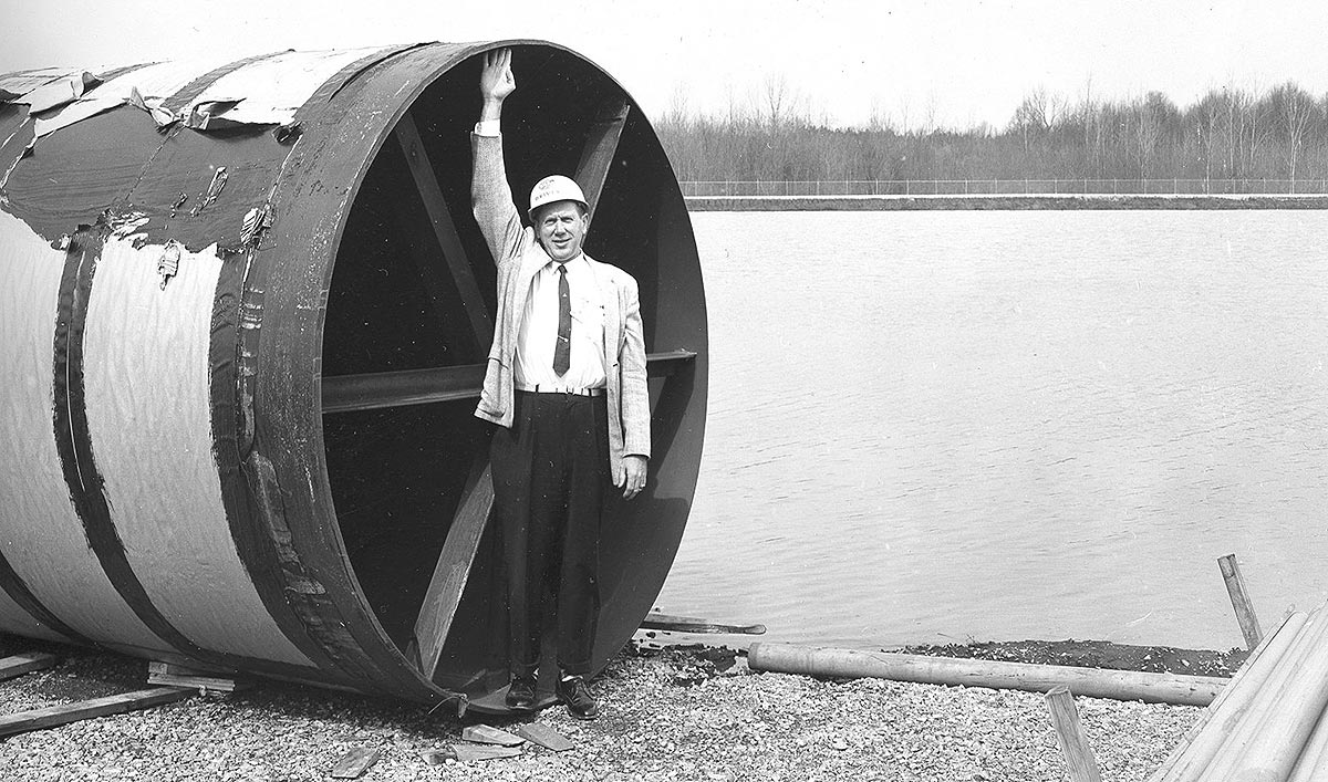 Description: A man in tie, sweater, and hardhat is standing in the mouth of a huge pipe. He smiles into the camera, and reaches his arm up so his fingers just touch the top of the pipe, several feet above his head. The pipe is lying on the grass in a construction site. Behind him is a canal.