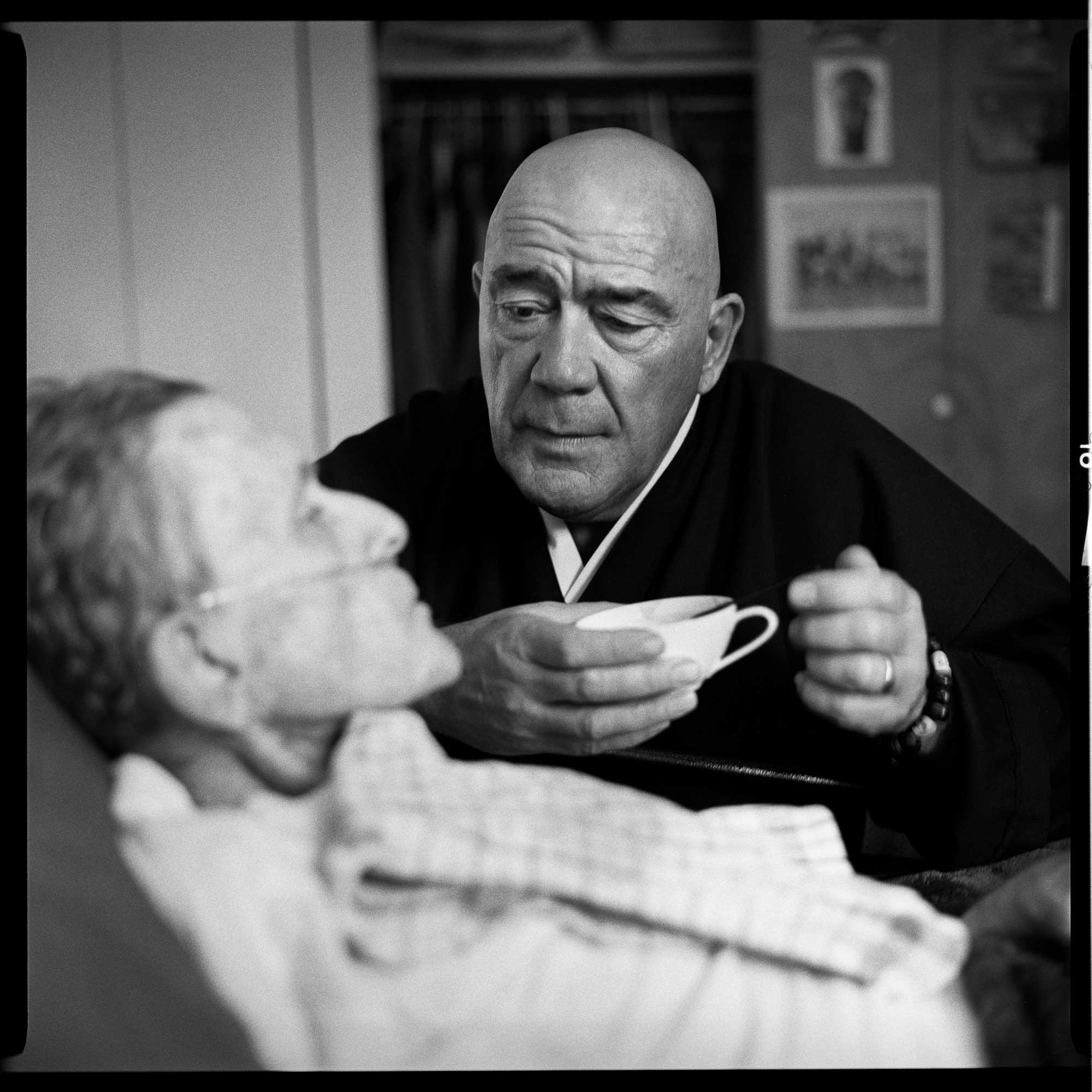 Black-and-white photo of Chodo sitting beside John. John lies in bed with an oxygen tube under his nose. Chodo holds a cup of soup and a spoon and looks at John's face.