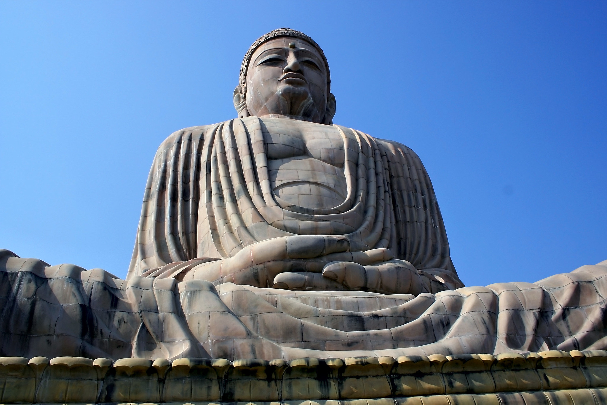 A color photograph of a huge stone Buddha in Bodh Gaya, India. The photo is taken from a low angle so the Buddha towers over the viewer. In the foreground, his hands are folded in his lap and they appear enlarged. His distant face is stern.