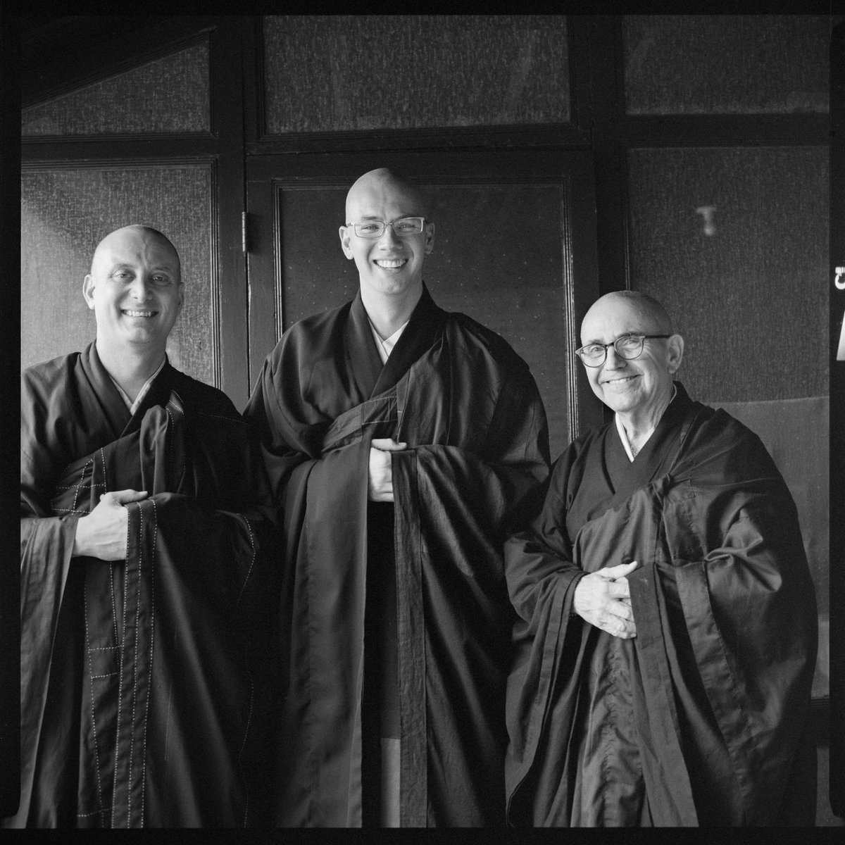 Image description: black and white photo of three men with shaved heads smiling into the camera, dressed in black robes.
