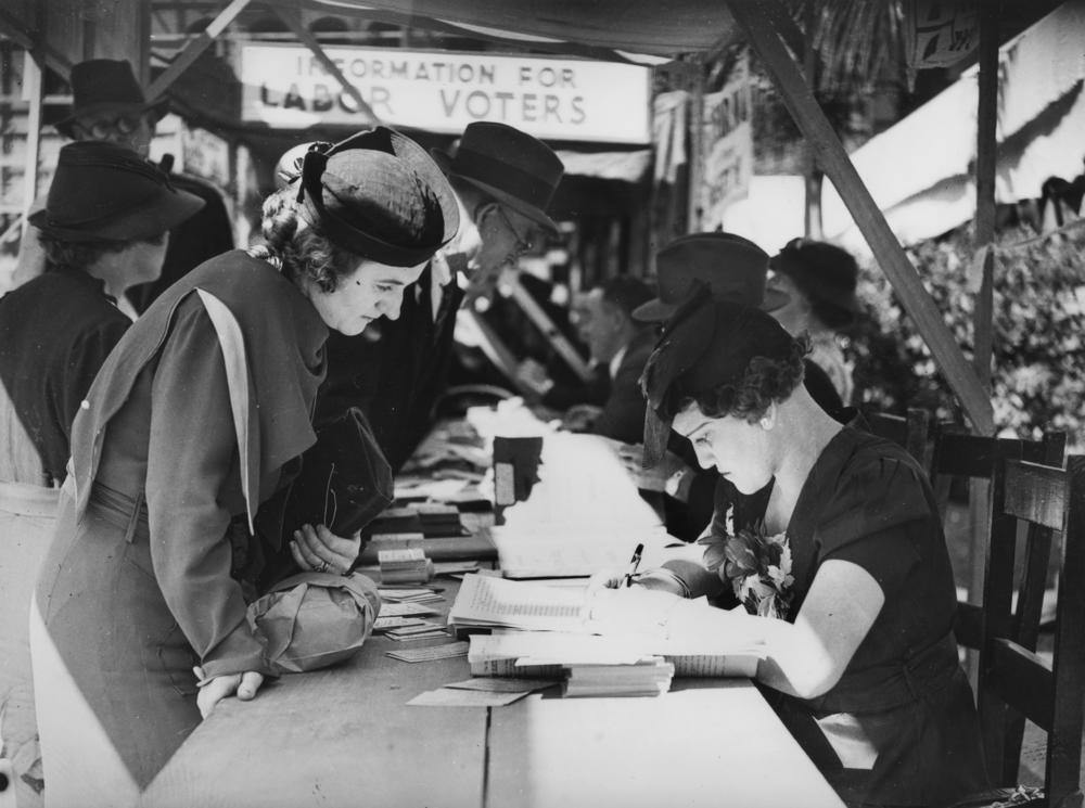 Black and white photograph of voters in 1930s-era British dress, standing lined up on one side of a wooden table, consulting with poll workers seated on the other side of the table and checking voter rolls.