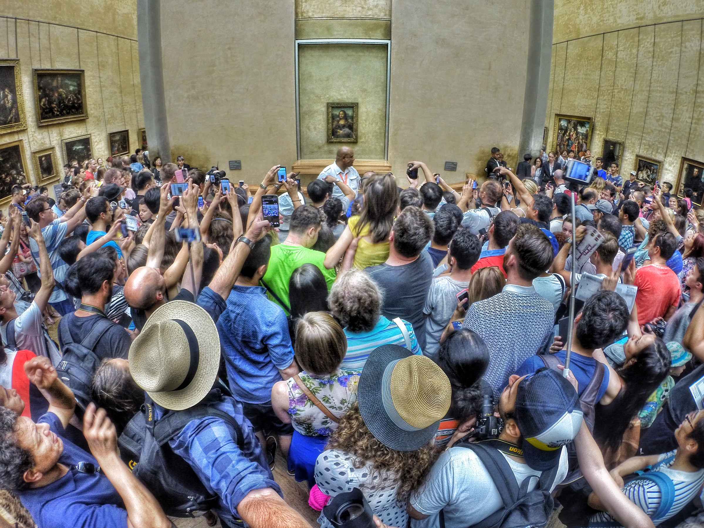 Color photo of a crowd at the Louvre pressing close to the Mona Lisa, which is displayed with a whole wall to itself. Most people in the crowd are holding up phones or cameras to photograph the painting. A security guard stands directly in front of the painting, keeping the crowd back.