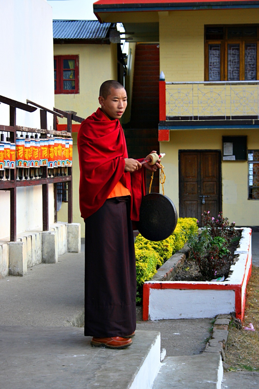 A young Tibetan monk in red robes holds a small black hand-gong and a striker, standing on a path in front of a row of prayer wheels at a monastery in Sikkim